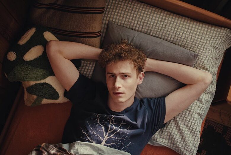 Pokazany od góry młody chłopak leżący w łóżku w zrelaksowanej pozie, patrzący wprost do obiektywu.