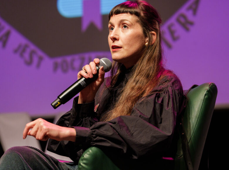 Zbliżenie na siedzą w fotelu kobietę, trzymającą w ręku mikrofon i mówiącą coś do niego. Za nią w tle kolorowa ściana.