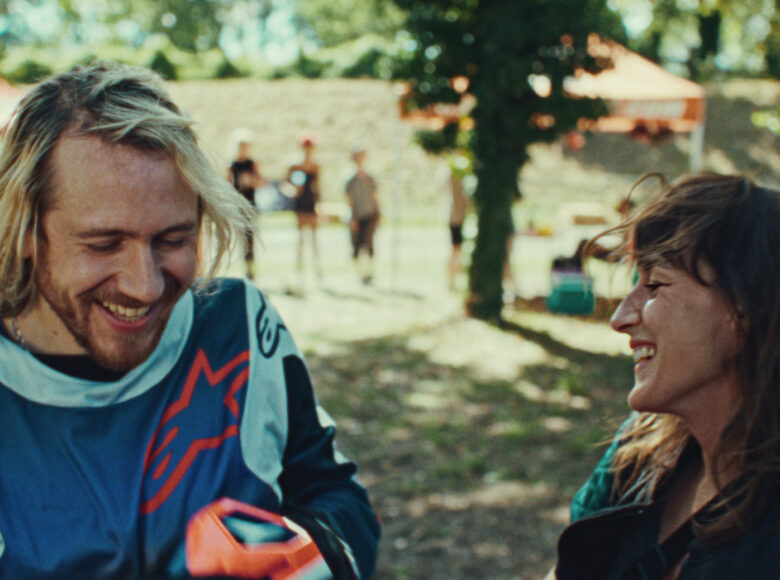 Zdjęcie z filmu. Ciepły dzień w parku. Na pierwszym planie kobieta i mężczyzna stoją naprzeciwko siebie i śmieją się. W tle stoi kilka innych osób, wokół rosną zielone drzewa i trawa.