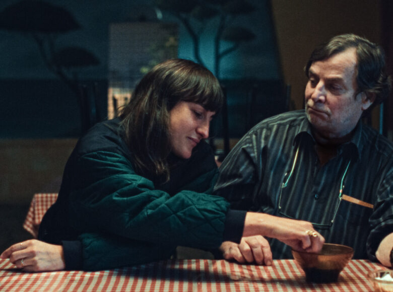 Wnętrze stołówki. Półmrok. Na pierwszym planie przy stole siedzą młoda kobieta i mężczyzna w średnim wieku. Kobieta ubrana jest w kurtkę ortalionową, mężczyzna w koszulę. Mężczyzna patrzy na kobietę. Kobieta patrzy na stojąca na stole małą miskę, sięgając do niej ręką. W tle puste stoły.
