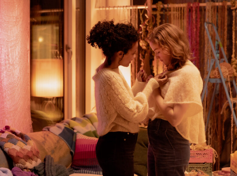 Wnętrze przytulnego pokoju oświetlonego ciepłym światłem lampy. Na pierwszym.planie stoją blisko siebie dwie dziewczyny. Zwrócone są do siebie twarzami. Jedna chwyta w palce kołnierz swetra, w który ubrana jest druga.