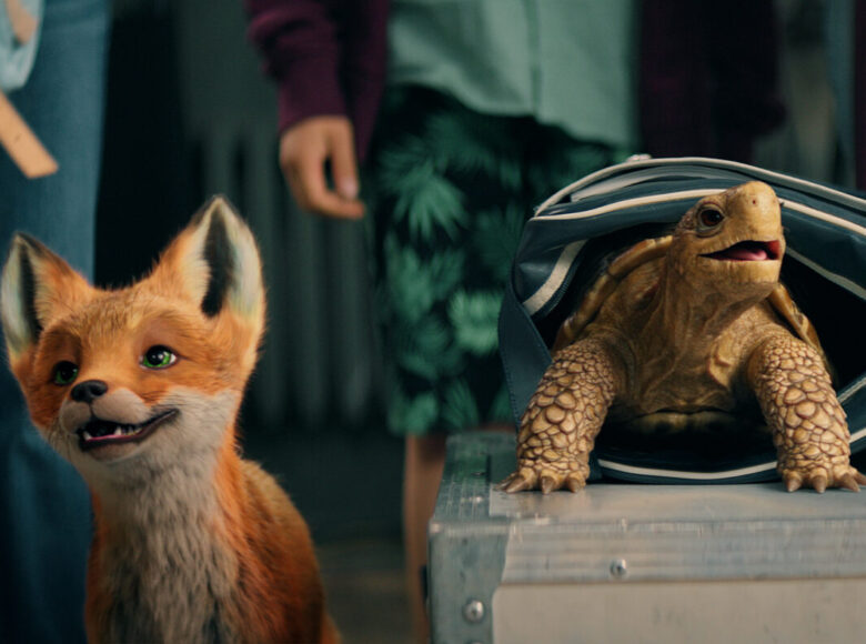 Kadr z filmu. Na pierwszym planie kukiełki przedstawiające zwierzątka - liska ora żółwia, który wychodzi z toby leżącej na kufrze. Na dalszym planie widać sylwetki kilkorga dzieci stojących obok siebie.