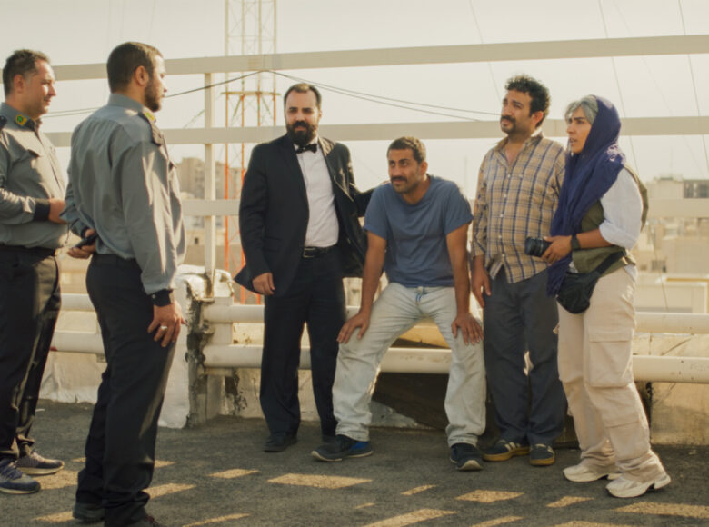 Letni, ciepły dzień. Na dachu budynku stoi kobieta i trzech mężczyzn. Rozmawiają z dwoma mężczyznami w uniformach służbowych, którzy wyglądają jak ochroniarze lub funkcjonariusze jakichś służb.