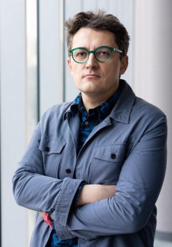 Zbliżenie na mężczyznę w średnim wieku, pokazanego od pasa w górę. Mężczyzna ubrany jest w szary żakiet. Nosi okulary w zielonych oprawkach. Znajduje się w jasnym, białym pomieszczeniu. Stoi na tle okna zasłoniętego białymi żaluzjami.