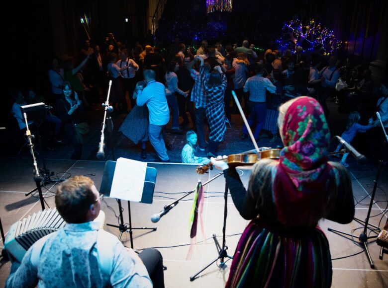 Duża sala. Na pierwszym planie widzimy od tyłu kobietę w ludowym stroju grającą na skrzypcach i chłopaka grającego na akordeonie. Na dalszym planie bardzo duża grupa ludzi tańczących w parach.