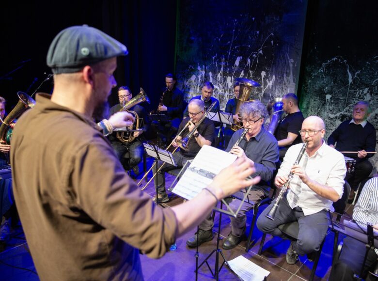 Na scenie grupa mężczyzn i kobiet grających na instrumentach dętych. Przed nimi stoi dyrygent.