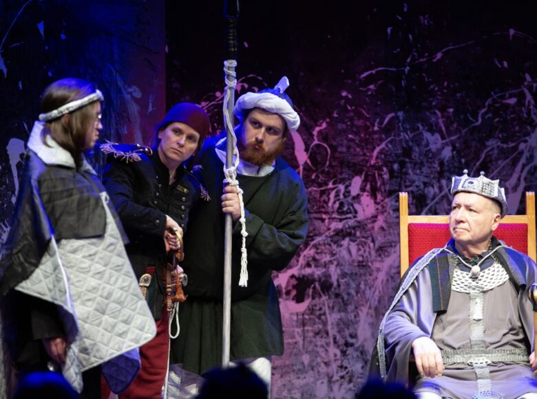 Przedstawienie teatralne. Na scenie siedzący na tronie aktor grający króla. Obok niego stoi trójka aktorów - kobieta w kostiumie nawiązującym do dziewiętnastowiecznego munduru wojskowego, mężczyzna w kostiumie nawiązującym do mody średniowiecznej i osoba w kostiumie złożonym z obszernych płacht, uniemożliwiającym określenie jej płci. Za aktorami kolorowa ściana z abstrakcyjnymi wzorami.