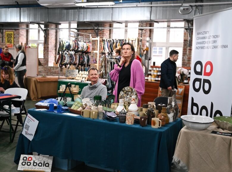 Wnętrze dużej hali, w której odbywają się targi rzemiosła. Na pierwszym planie stoisko z ceramicznymi naczyniami. W tle inne stoiska z przetworami kulinarnymi, modą czy z plakatami artystycznymi. Przy stoiskach siedzą i stoją osoby sprzedające i oglądające.