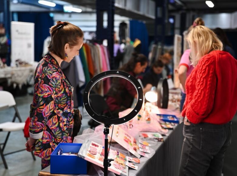 Wnętrze dużej hali, w której odbywają się targi rzemiosła. Na pierwszym planie stoisko z biżuterią. W tle inne stoiska, na przykład z modą. Przy stoiskach siedzą i stoją osoby sprzedające i oglądające.