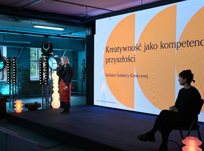 Konferencja. Na scenie dwie kobiety: jedna stoi i trzyma w ręku mikrofon mówiąc coś do niego. Druga siedzi na krześle, parząc na pierwszą kobietę. Za nimi z tyłu sceny duży napis: "Kreatywność jako kompetencja przyszłości. Inkubator Dzielnicy Kreatywnej".