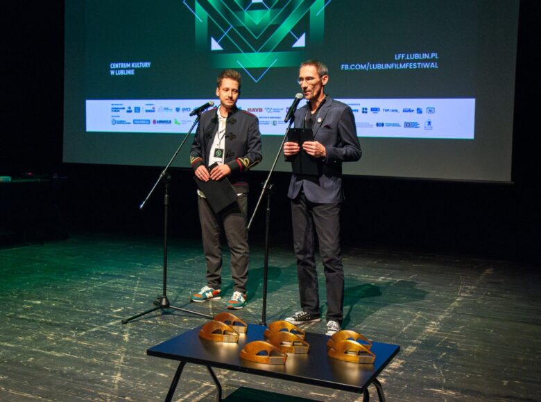 Na scenie przy mikrofonach stoją dwaj mężczyźni. Przed nimi na stoliku stoi siedem statuetek. Za nimi z tyłu sceny wisi duże logo Lubelskiego Festiwalu Filmowego.