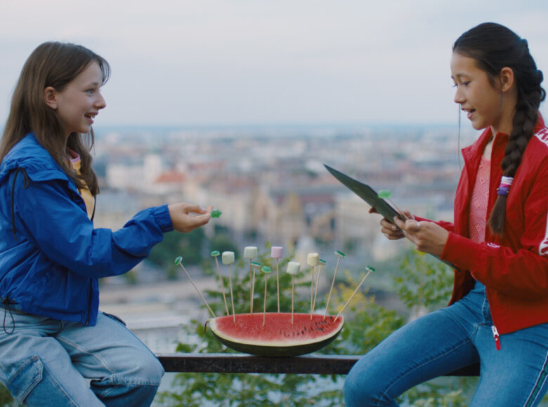 Kadr z filmu. Na pierwszym planie dwie kilkunastoletnie dziewczynki siedzące naprzeciwko siebie na balustradzie tarasu. Między dziewczynkami, także na balustradzie leży ćwiartka arbuza, w którą powtykane są słodycze nadziane na patyki. W tle widać szeroki widok z lotu ptaka na miasto.