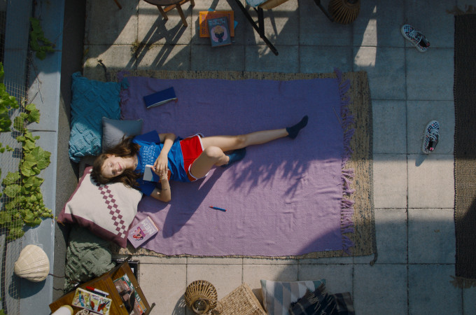 Na tarasie pokazanym od góry leży na kocu dziewczyna w letnich ubraniach. W dłoniach trzyma książkę.