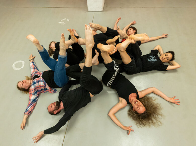 W sali do ćwiczeń gimnastycznych na podłodze leży obok siebie kilka osób, unosząc nogi do góry.