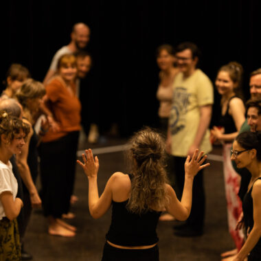 W ciemnawej sali kilka osób, kobiet i mężczyzn, stoi w kręgu. Wszyscy są ubrani w stroje do ćwiczeń gimnastycznych.
