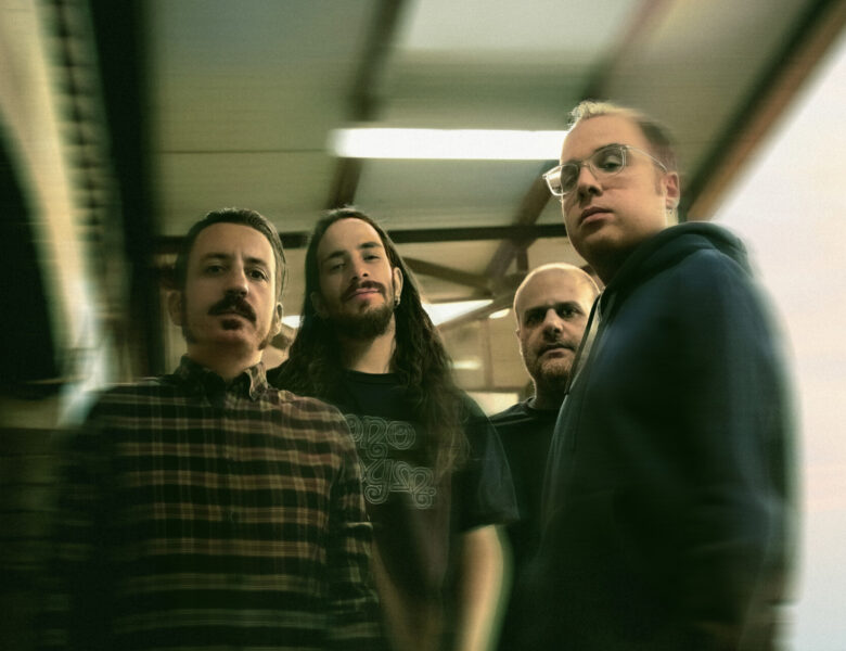 W pomieszczeniu stoją blisko siebie czterej mężczyźni, pozując do fotografii. Patrzą prosto na nas.