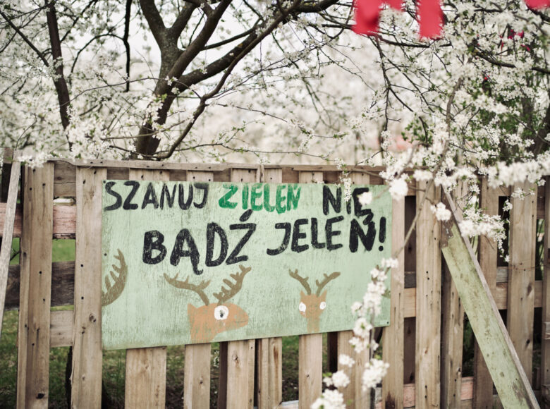 Na drewnianym płocie wisi tablica z odręcznie namalowanym napisem: "Szanuj zieleń nie bądź jeleń!". Za płotem rosną drzewka owocowe, kwitnące na biało.