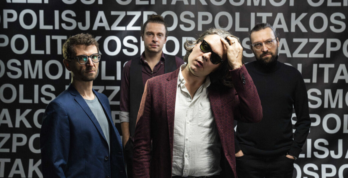 Czterej mężczyźni w średnim wieku pozują do fotografii na tle czarnej ściany pokrytej białymi napisami.