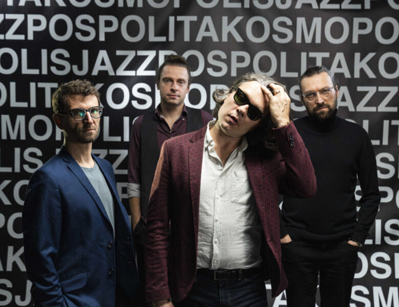 Czterej mężczyźni w średnim wieku pozują do fotografii na tle czarnej ściany pokrytej białymi napisami.