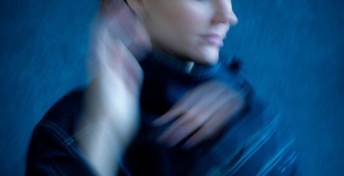 Na fotografii rozmyty portret kobiety unoszącej dłonie ku twarzy. Za kobietą niebieskie tło.