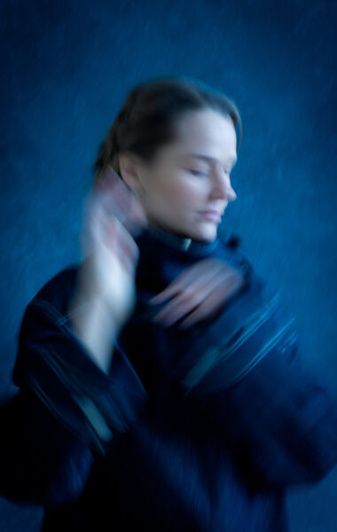 Na fotografii rozmyty portret kobiety unoszącej dłonie ku twarzy. Za kobietą niebieskie tło.