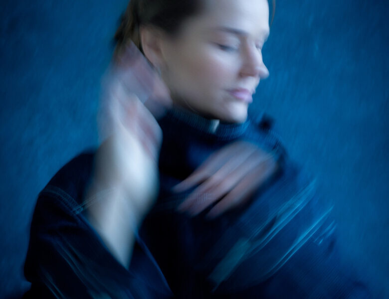 Na fotografii rozmyty portret kobiety unoszącej dłonie ku twarzy. Za kobietą niebieskie tło.