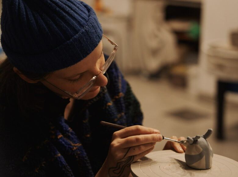 Przy stole w pracowni siedzi kobieta i maluje wzory na ceramicznej figurce przedstawiającej ptaszka.