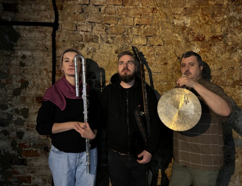 W pomieszczeniu na tle ceglanej ściany stoją, pozując do fotografii, młoda kobieta i dwóch młodych mężczyzn. Wszyscy trzymają w rękach instrumenty muzyczne.