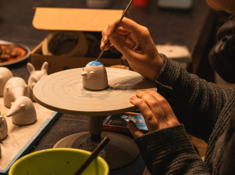 Przy stole w pracowni siedzi kobieta i maluje wzory na ceramicznej figurce przedstawiającej ptaszka.