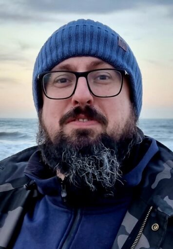 Zbliżenie na twarz i ramiona mężczyzny stojącego na brzegu morza. Mężczyzna patrzy prosto w obiektyw. Ubrany jest w ciepłą kurtkę i czapkę. Ma wąsy, brodę i okulary.