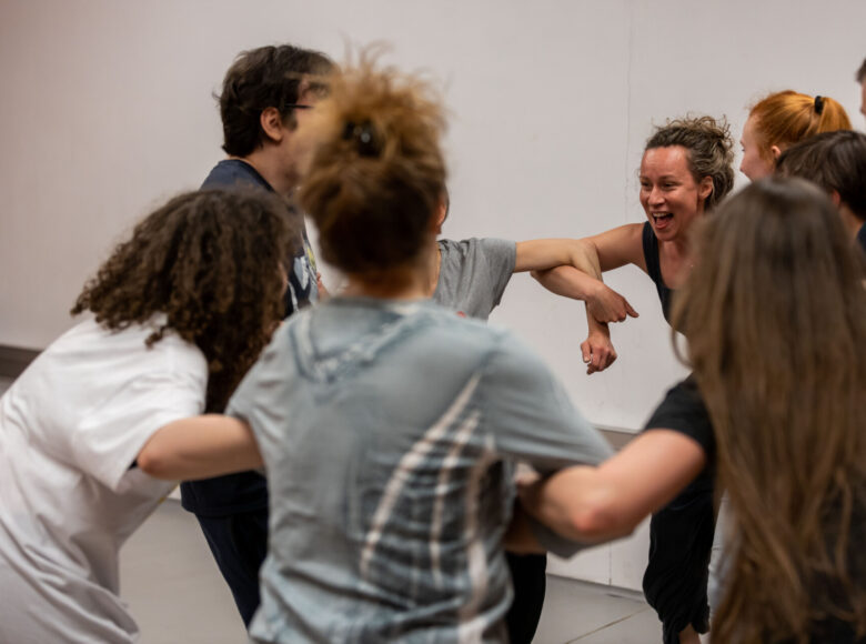 W sali kilka osób porusza się w kręgu, trzymając jedna drugą pod rękę. Wszystkie ubrane są w stroje do ćwiczeń gimnastycznych.