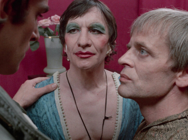 Zbliżenie na trzech mężczyzn stojących blisko siebie w kręgu. Jeden z nich ma kobiecy makijaż i głęboki dekolt. W tle czerwona ściana i bukiet róż w wazonie na szafce pod ścianą.