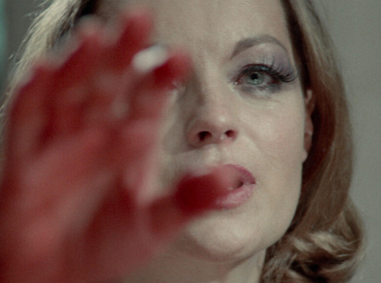 Zbliżenie na twarz kobiety. Kobieta ma jasne, rozpuszczone włosy do ramion, pomalowane oczy i pomadkę na ustach. Wyciąga do przodu dłoń, częściowo zasłaniając swoją twarz.