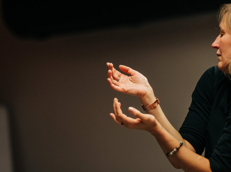 W ciemnym pomieszczeniu na pierwszym planie stoi kobieta z zamkniętymi oczami, unosząc do góry dłonie. Za kobietą szare, ciemne tło.