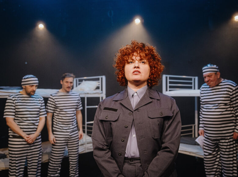 Spektakl teatralny. Na scenie na pierwszym planie stoi kobieta w szarym mundurze. Za nią, przy dwóch piętrowych łóżkach stoją trzej mężczyźni w pasiastych koszulach, spodniach i czapkach.