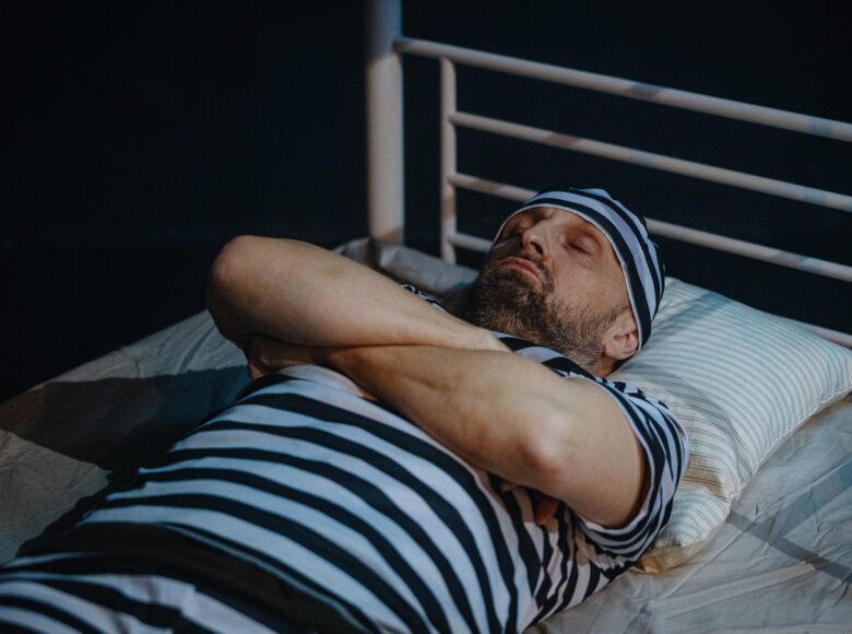 Spektakl teatralny. Na metalowym łóżku leży mężczyzna w pasiastej koszuli, spodniach i czapce. Ma zamknięte oczy.