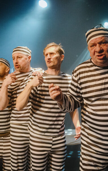 Spektakl teatralny. Na scenie stoją obok siebie czterej mężczyźni w pasiakach.