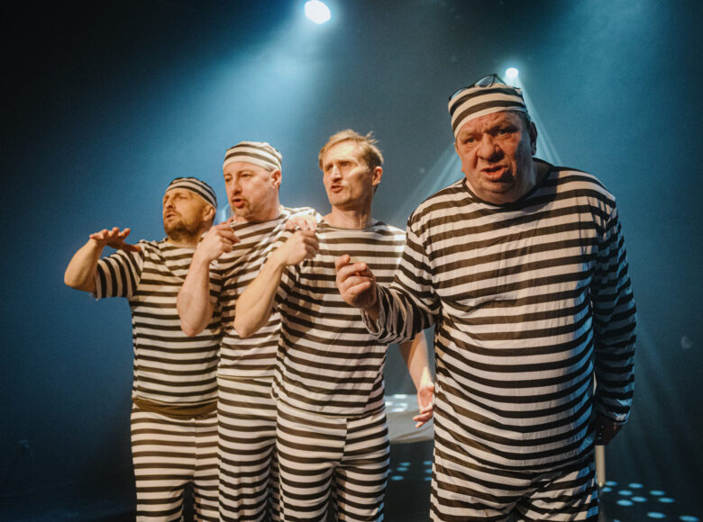 Spektakl teatralny. Na scenie stoją obok siebie czterej mężczyźni w pasiastych koszulach, spodniach i czapkach.