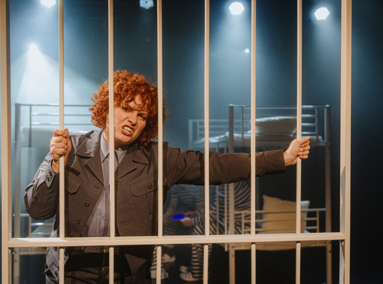 Spektakl teatralny. Na scenie stoi za kratą kobieta w szarym mundurze. Za kobietą w tle stoją dwa łózka piętrowe. Na łóżkach siedzą trzej mężczyźni w pasiakach.