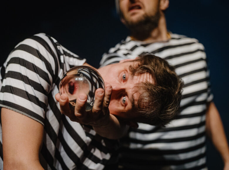 Spektakl teatralny. Na scenie stoją dwaj mężczyźni w pasiakach. Pierwszy pochyla się i patrzy z uwagą w szklaną kulę, którą trzyma w dłoni. Drugi stoi za nim i patrzy w górę z zaciekawieniem.