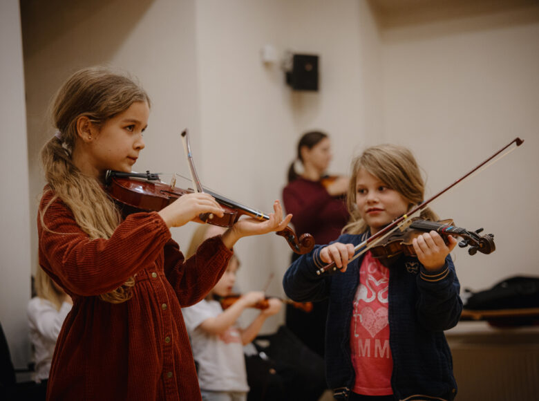 W dużym pomieszczeniu kilka dziewczynek gra na skrzypcach.