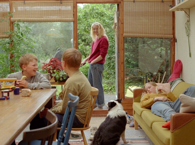 W pokoju przy stole siedzi dwóch roześmianych chłopców, obok na sofie leży dziewczynka, a na podłodze siedzi pies. Obok dzieci, w drzwiach prowadzących do ogrodu stoi uśmiechnięta kobieta. Kobieta rozmawia z chłopcami.