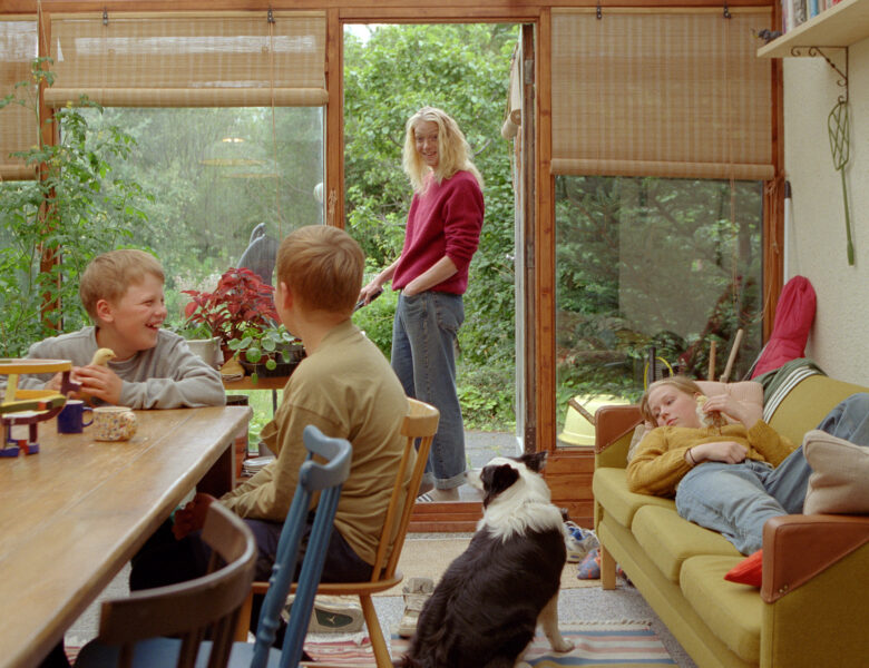 W pokoju przy stole siedzi dwóch roześmianych chłopców, obok na sofie leży dziewczynka, a na podłodze siedzi pies. Obok dzieci, w drzwiach prowadzących do ogrodu stoi uśmiechnięta kobieta. Kobieta rozmawia z chłopcami.