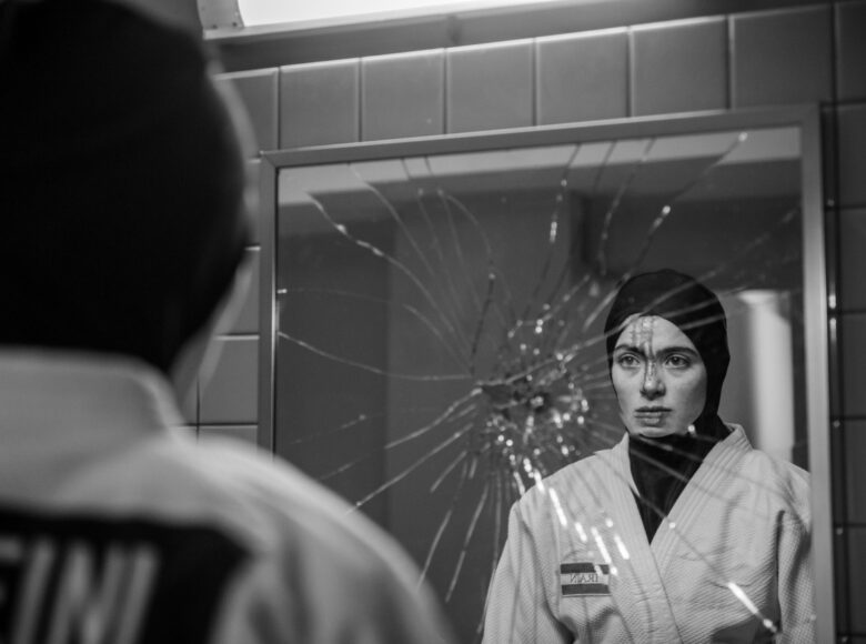 Fotografia czarno biała. W łazience dziewczyna w muzułmańskiej chuście na głowie patrzy na swoje odbicie w rozbitym lustrze.