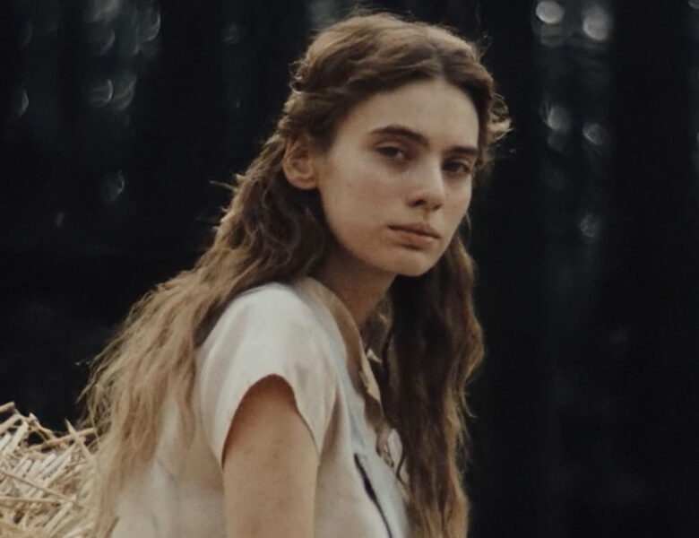 Zbliżenie na dziewczynę z długimi, rozpuszczonymi włosami i ubraną w letni strój. Dziewczyna patrzy na widza. Za nią ciemne tło z delikatnymi punktami świateł.