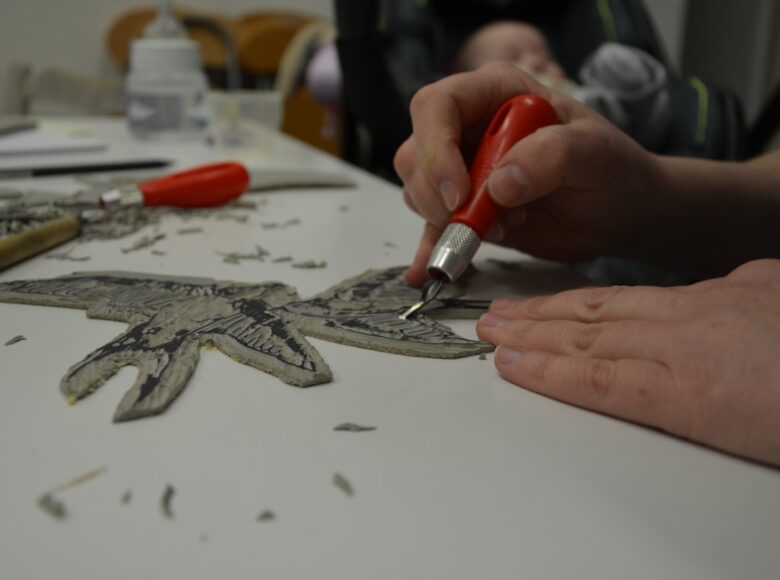 Zbliżenie na dłonie osoby wycinającej dłutem wzór w niewielkim kawałku linoleum. Osoba ta tworzy w ten sposób matrycę do odbitek graficznych.