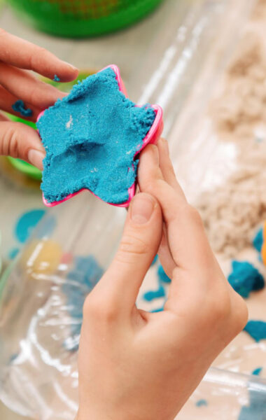 Zbliżenie na dłonie młodej osoby, wkładającej masę modelarską do foremki w kształcie gwiazdki. Poniżej widać blat stołu, na którym leżą inne plastikowe foremki i kawałki masy modelarskiej.