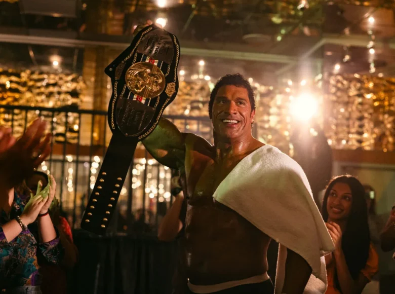 Wnętrze w budynku publicznym. Umięśniony mężczyzna z nagim torsem unosi do góry trofeum sportowe. Dookoła stoi kilka kobiet, które patrzą na mężczyznę i biją brawo.