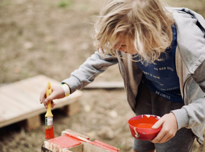 W plenerze na trawniku kilkuletni chłopiec maluje drewnianą skrzynkę czerwoną farbą.