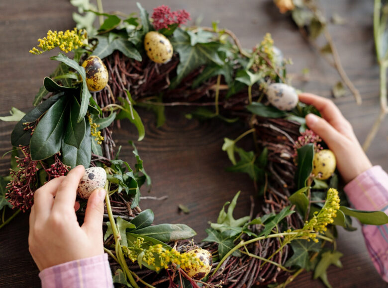Zbliżenie na ręce osoby, która umieszcza jajeczko przepiórki na wianku z gałązek i liści, leżącym na stole.